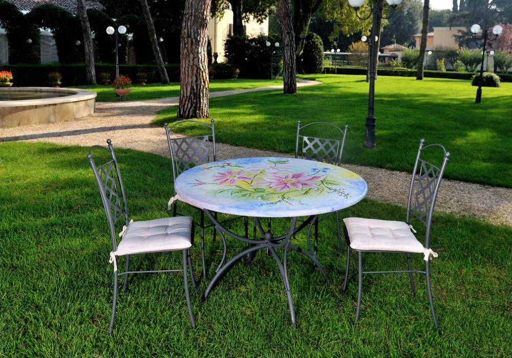 Tavolo rotondo dipinto con fiori rosa e sedie in ferro battuto con cuscini bianchi su prato verde, giardino.