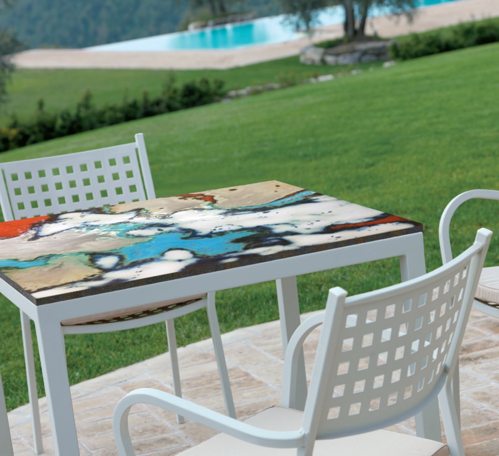 Tavolo da esterno bianco con piano artistico astratto, sedie abbinate, prato verde e piscina sullo sfondo.