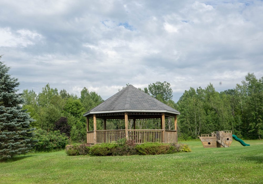 Gazebo in legno con tetto scuro in un prato verde, circondato da alberi.