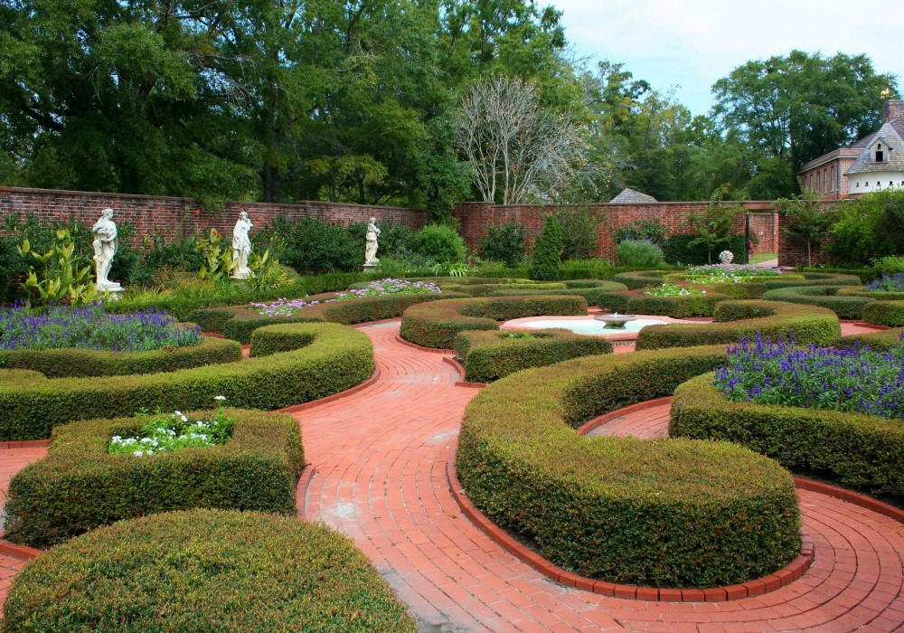 Giardino formale con siepi curve, vialetti in mattoni rossi, statue bianche e alberi verdi sotto un cielo nuvoloso.