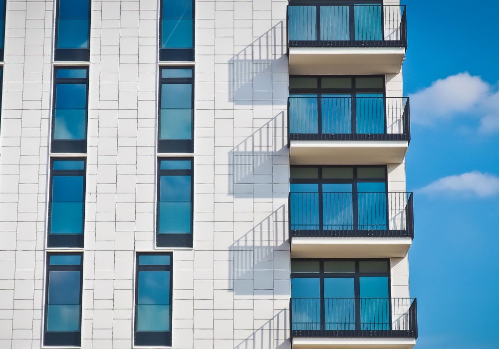 Facciata moderna di un edificio bianco con finestre blu e balconi neri, cielo azzurro con nuvole.