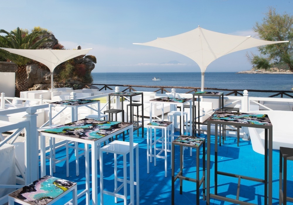 Terrazza bar sul mare con tavoli alti, sgabelli e ombrelloni bianchi, pavimento blu.