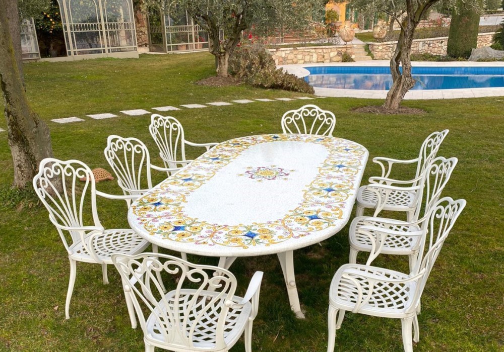 Tavolo ovale da giardino in mosaico e sedie bianche in ferro battuto su prato verde con piscina e villa sullo sfondo.