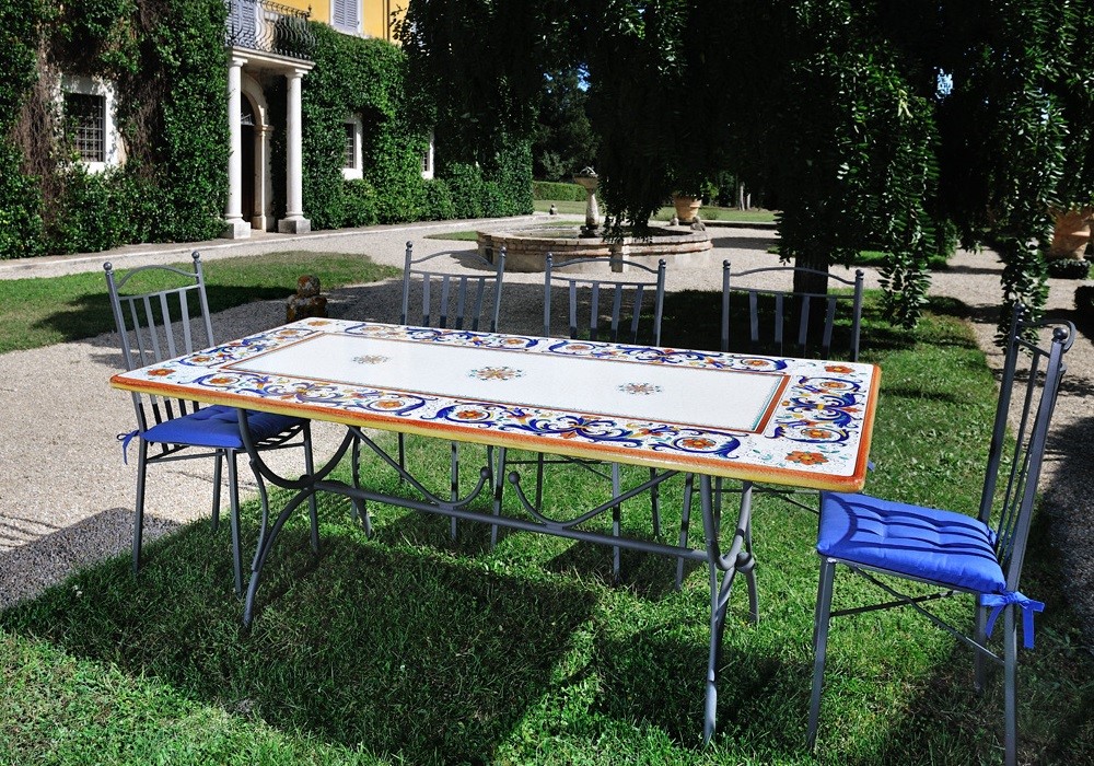 Tavolo rettangolare in ceramica decorata con sedie in ferro battuto e cuscini blu in un giardino italiano.