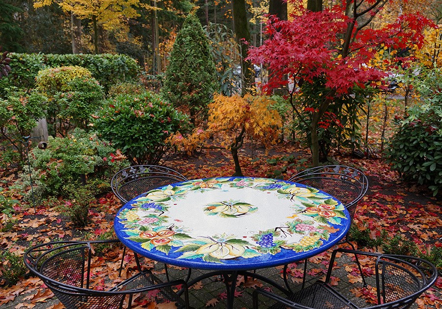 Tavolo da giardino in ferro battuto con piano decorato, circondato da alberi e arbusti con foglie autunnali.