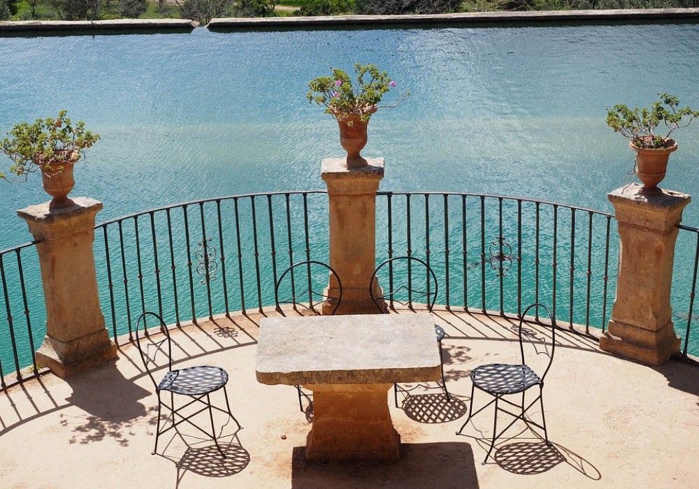 Balcone con tavolo e sedie in ferro battuto, vasi di fiori e ringhiera che si affaccia su una piscina blu.