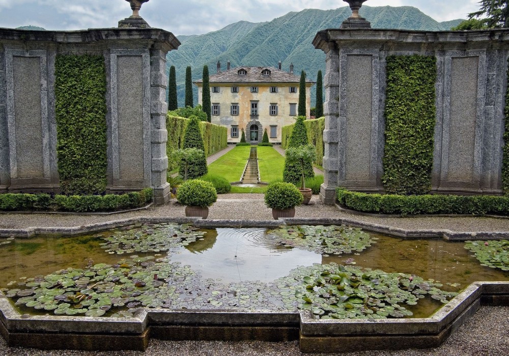 Giardino all'italiana con villa storica, laghetto di ninfee, siepi e montagne sullo sfondo.