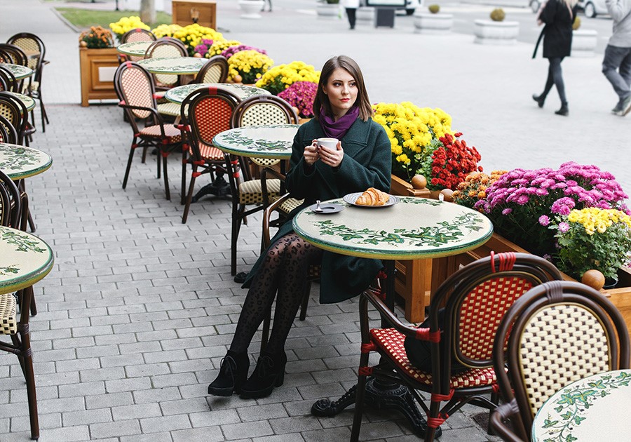 Donna in cappotto verde e sciarpa viola sorseggia caffè al tavolo di un caffè all'aperto con fiori colorati.