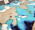 Tavolo in resina epossidica con design astratto che ricorda una mappa, toni di blu, turchese, bianco e sabbia.