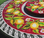 Tavolo rotondo in ceramica decorato a mano con limoni gialli, foglie verdi e fiori bianchi su sfondo rosso.