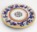 Piatto decorato in ceramica di Caltagirone con motivi floreali blu, arancioni e gialli su sfondo bianco.